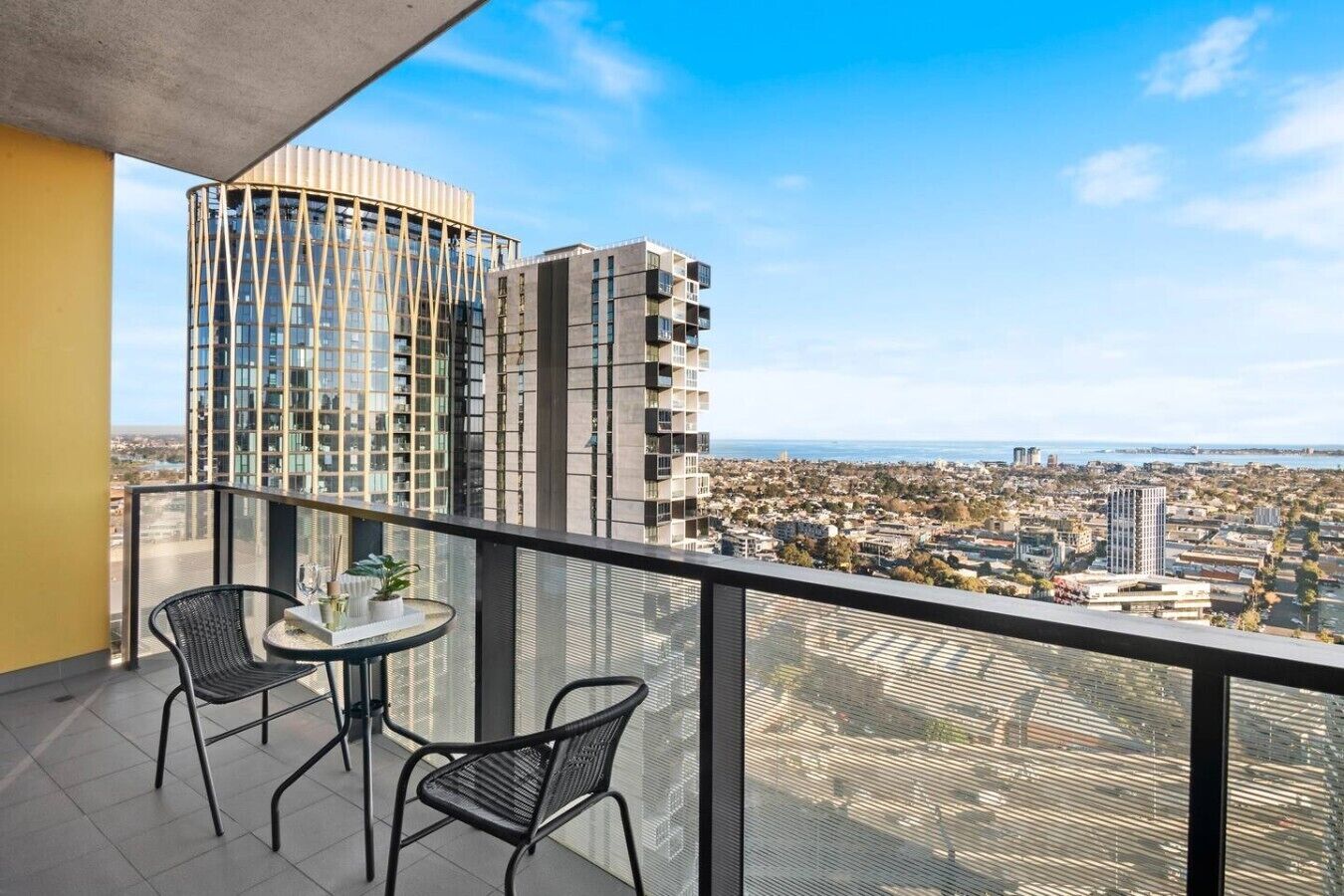 A balcony with two chairs and a small table, overlooking a cityscape with modern buildings and a clear sky.