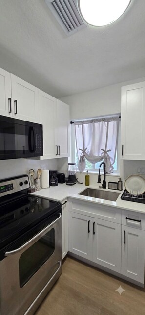 A modern kitchen with white cabinets, a black microwave, a stove, a sink with a window above, and a window with curtains. - Moderno Apto 2 Hab  Cerca Design District & MIA (MIAMI)