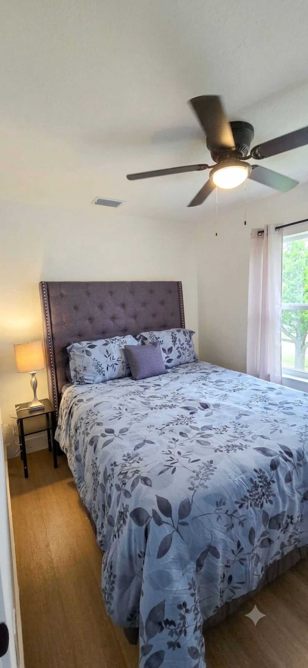 Un dormitorio con cama, ventilador de techo, mesita de noche y ventana.