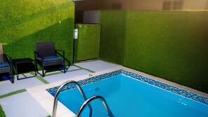 Ein Pool mit Fliesenrand, umgeben von Wänden aus Rasengittermatten, und zwei Korbstühle mit einem kleinen Tisch.