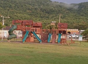 Ein Spielplatz mit Rutschen und Klettergerüsten, umgeben von einer Rasenfläche und einer hügeligen Landschaft.