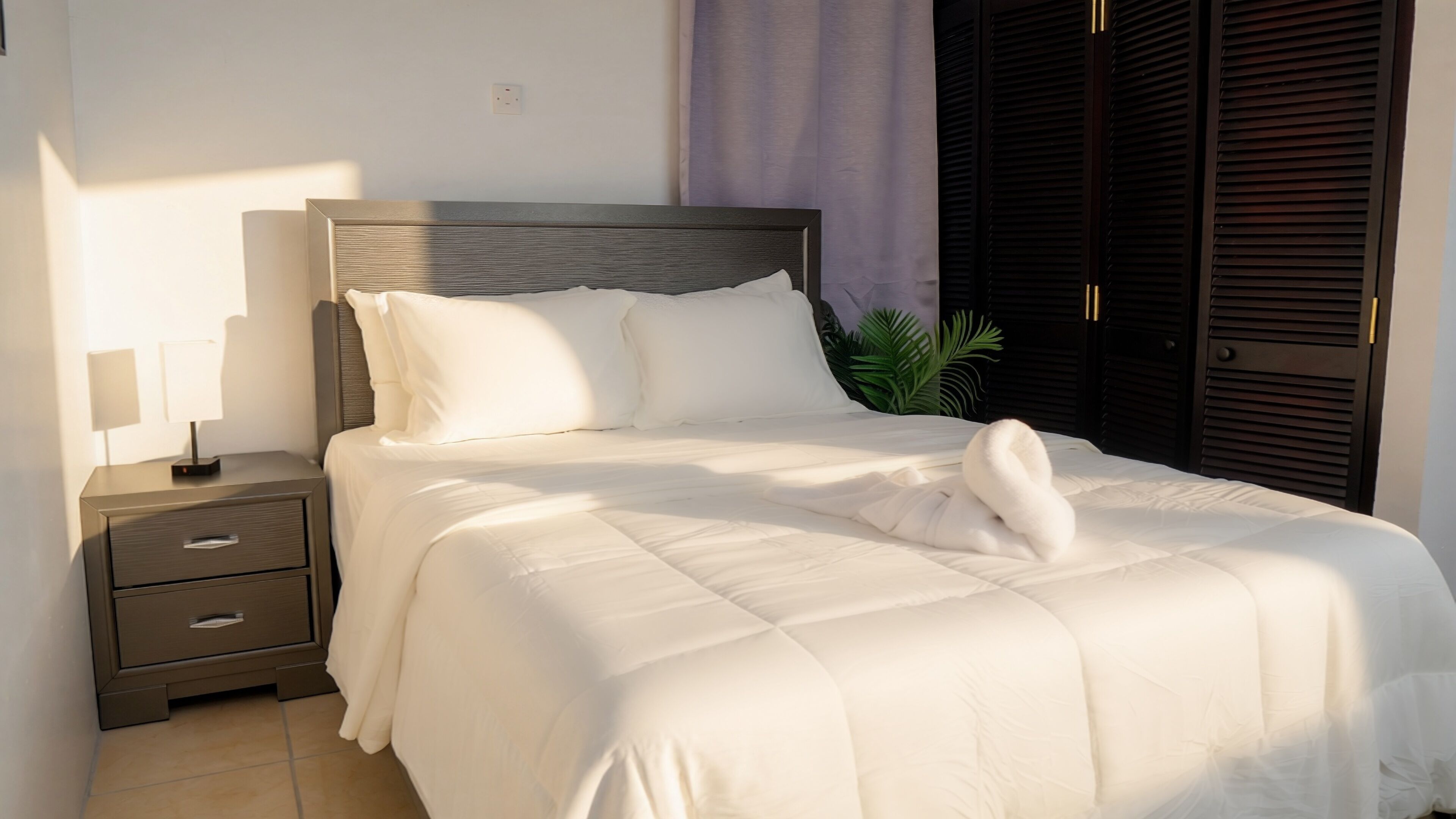 Una cama bien tendida con sábanas blancas y una toalla doblada, una mesita de noche con una lámpara y un armario de color oscuro.