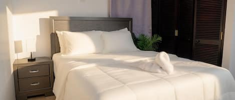 Una cama bien tendida con sábanas blancas y una toalla doblada, una mesita de noche con una lámpara y un armario de color oscuro.