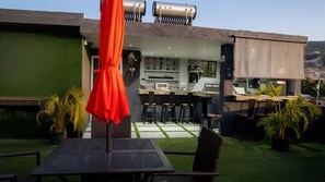 Eine Terrasse im Freien mit Tisch und Stühlen, einem roten Sonnenschirm und einem Grillbereich mit Blick auf die Landschaft.
