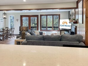 Ein modernes Wohnzimmer mit einem grauen Sofa, einem Esstisch mit Stühlen und großen Fenstern, die einen Blick auf Bäume bieten.