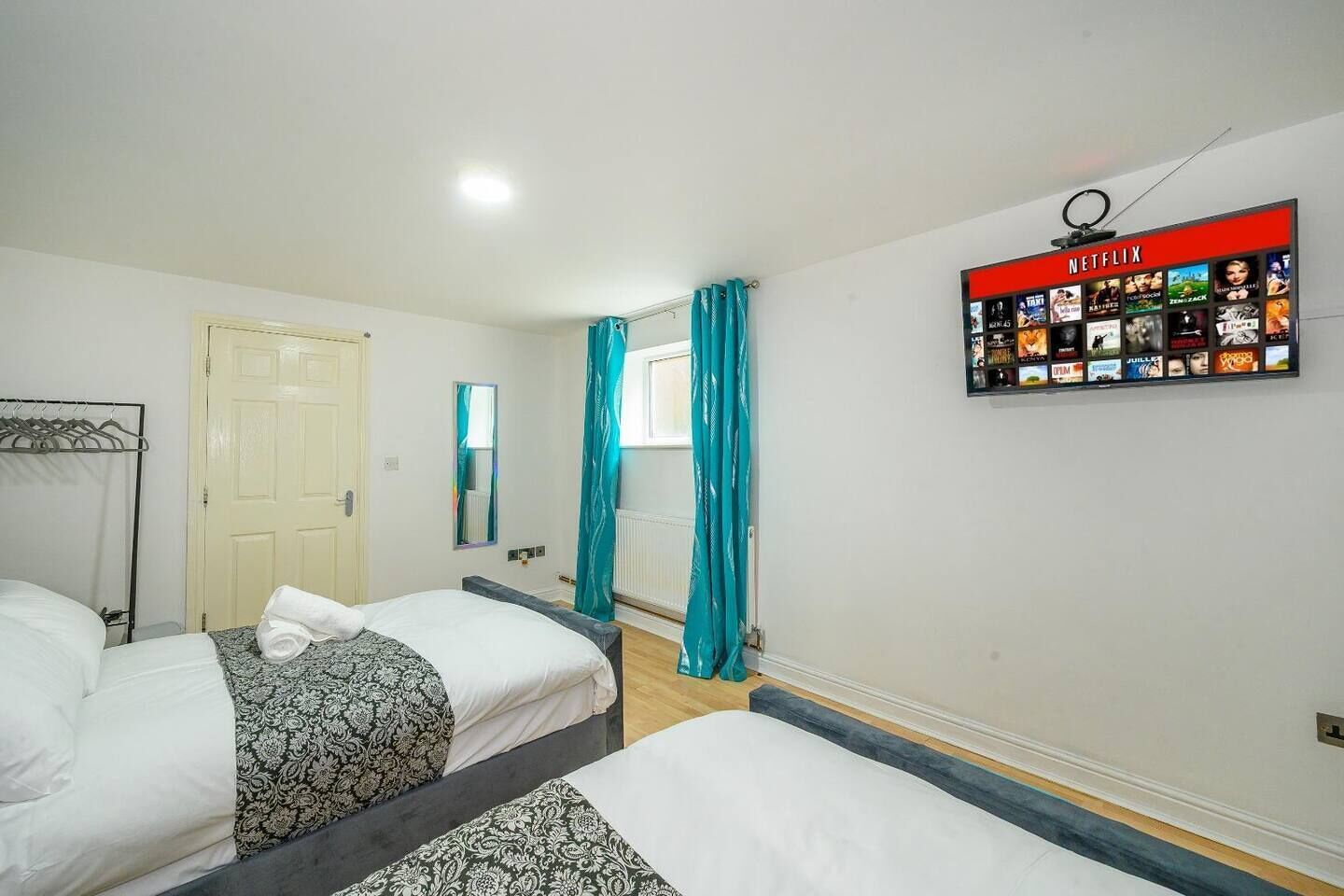 A hotel room with two beds, a wardrobe, a mirror, a window with curtains, and a wall-mounted Netflix sign.
