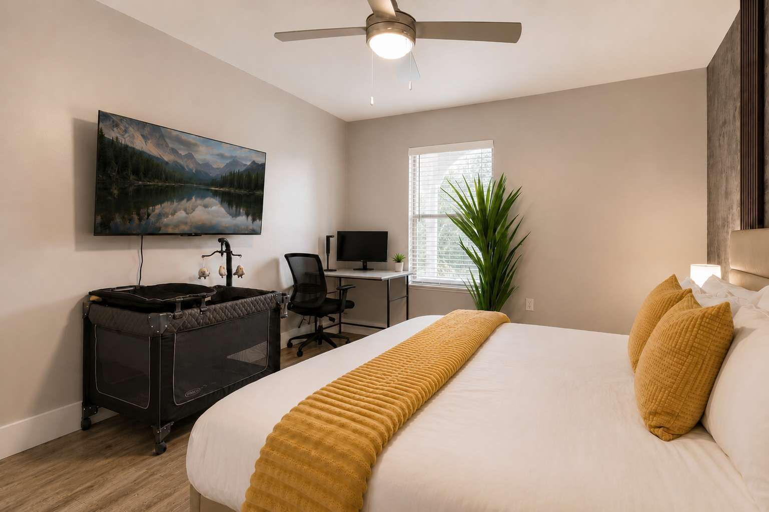 A hotel room with a bed, a desk with a computer, a television mounted on the wall, and a ceiling fan.