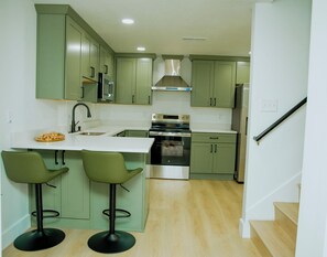 A kitchen with green cabinets, a stainless steel oven, and two bar stools. - Brand New 2BR Basement Suite +1 King Bed & Private Entrance (South Jordan)