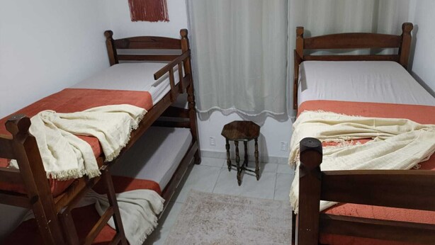 Una habitación con dos literas de madera, un taburete pequeño, un adorno de macramé en la pared y una ventana con cortinas blancas.