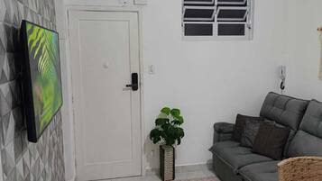 Um quarto com porta branca, ventilador de teto, sofá cinza, mesa pequena e uma planta.