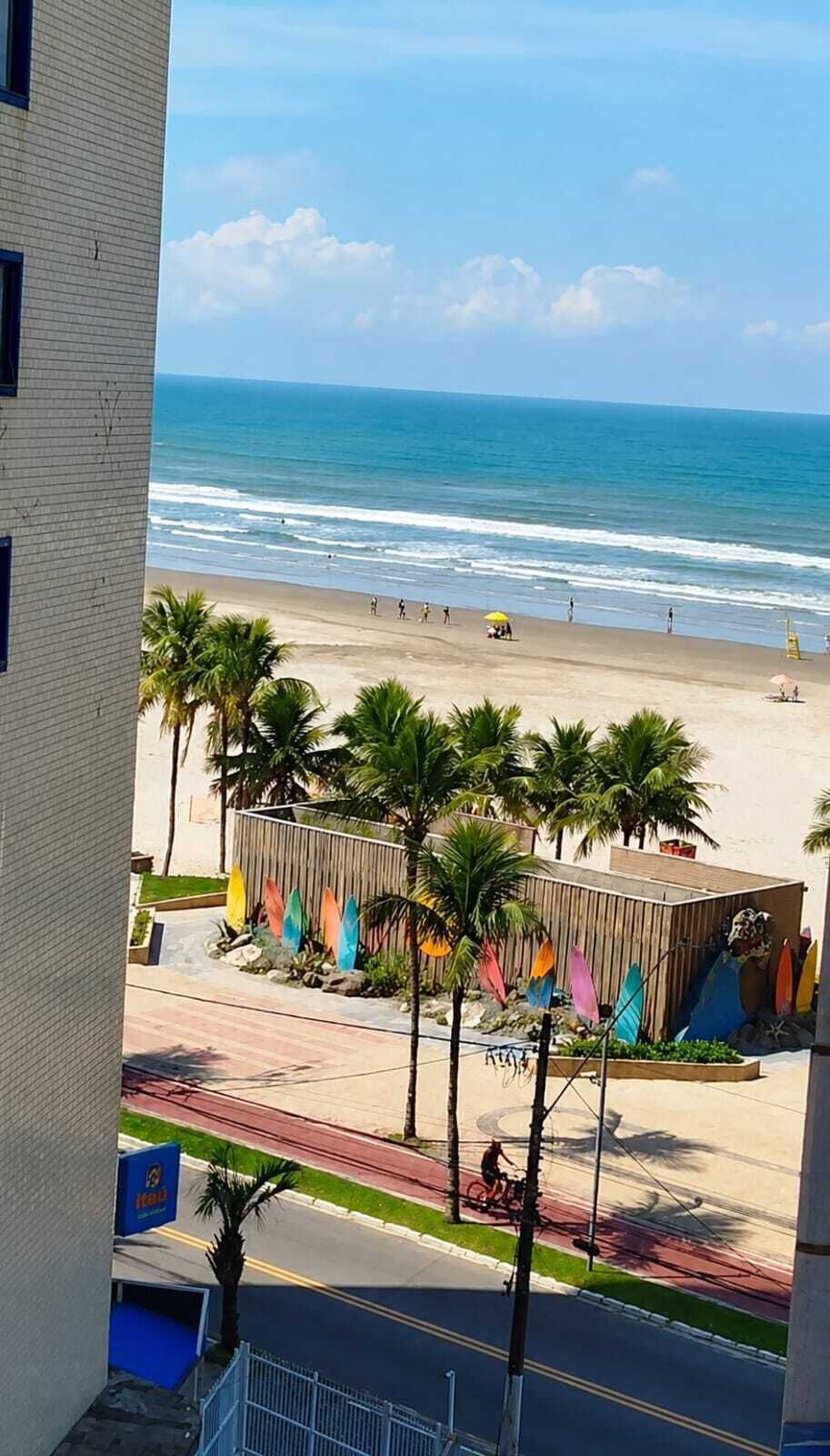 Une vue sur la plage depuis un immeuble, avec un ciel dégagé, des palmiers et une plage de sable où se trouvent des gens et un poste de surveillance des sauveteurs.