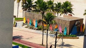 Vista de una playa desde un edificio, con cielo despejado, palmeras y una playa arenosa con gente y un puesto de salvavidas.
