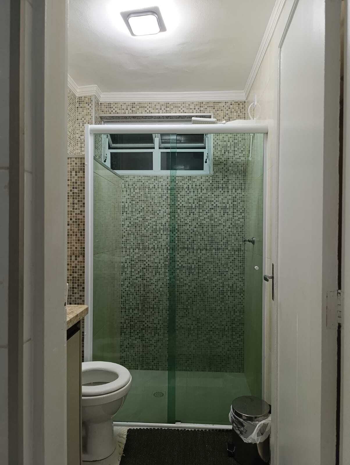 Uma casa de banho com chuveiro envolto em vidro, sanita e um pequeno contentor de lixo.