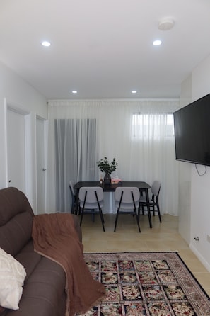 A living room with a brown sofa, a dining table with chairs, a flat-screen TV mounted on the wall, and a patterned rug on the floor. - New Modern Family Retreat 3BR Near Parramatta (Northmead)