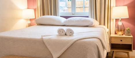 A neatly made bed with two rolled towels, a bedside table with a lamp, and a view of the outside through a window.