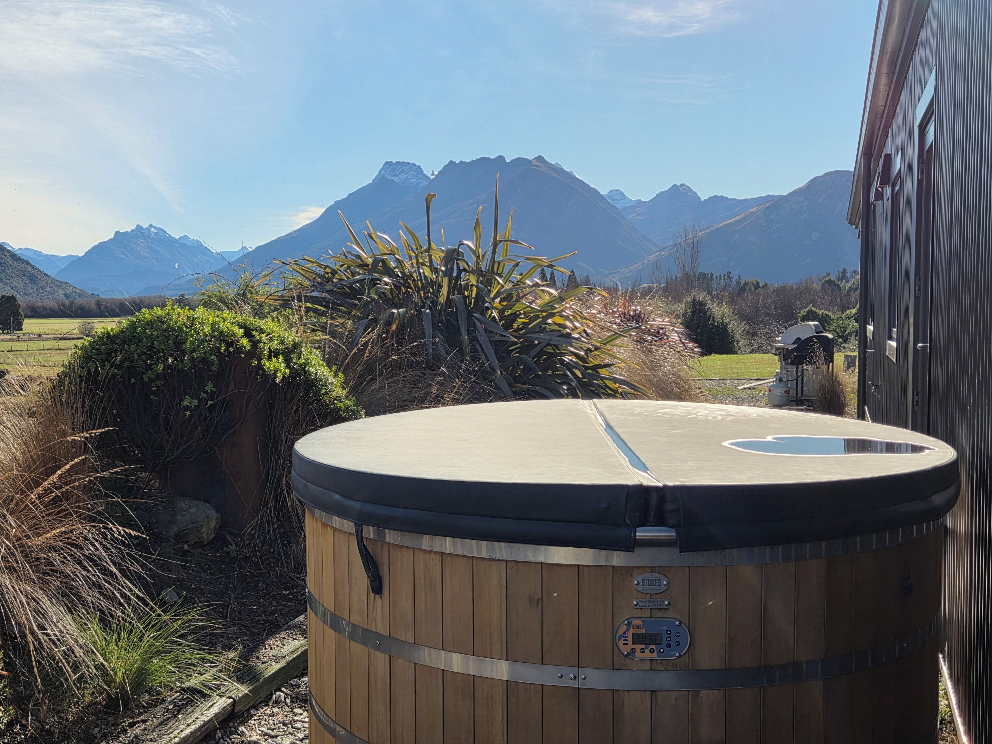 A wooden barrel-shaped hot tub with a digital control panel, surrounded by natural vegetation and mountains in the background.