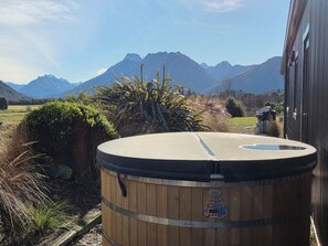 A wooden barrel-shaped hot tub with a digital control panel, surrounded by natural vegetation and mountains in the background.