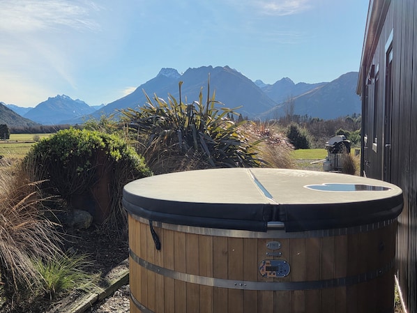 A wooden barrel-shaped hot tub with a digital control panel, surrounded by natural vegetation and mountains in the background.