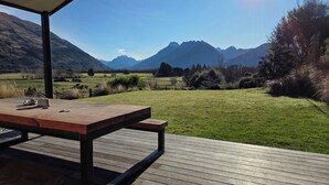 An outdoor wooden deck with a table and chairs, overlooking a grassy field and mountains. - Alpine Home Escape | Hot Tub & Mountain Views | Glenorchy (Glenorchy)