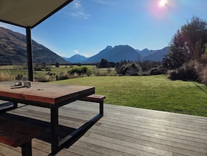Een terras van hout buiten, met een tafel en stoelen, met uitzicht op een grasveld en bergen.