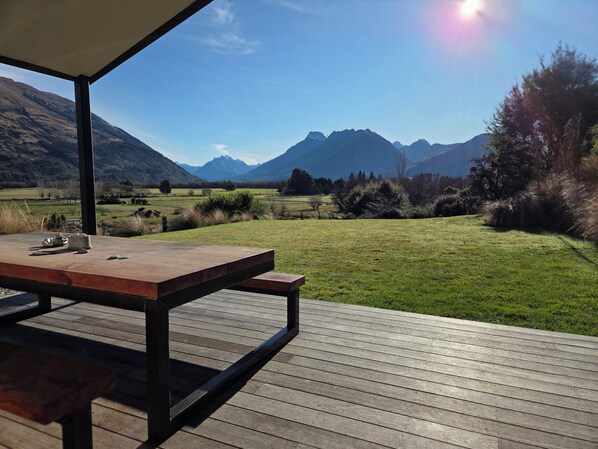 An outdoor wooden deck with a table and chairs, overlooking a grassy field and mountains.