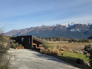 A modern building with a gravel driveway, surrounded by a grassy field and mountains in the background.