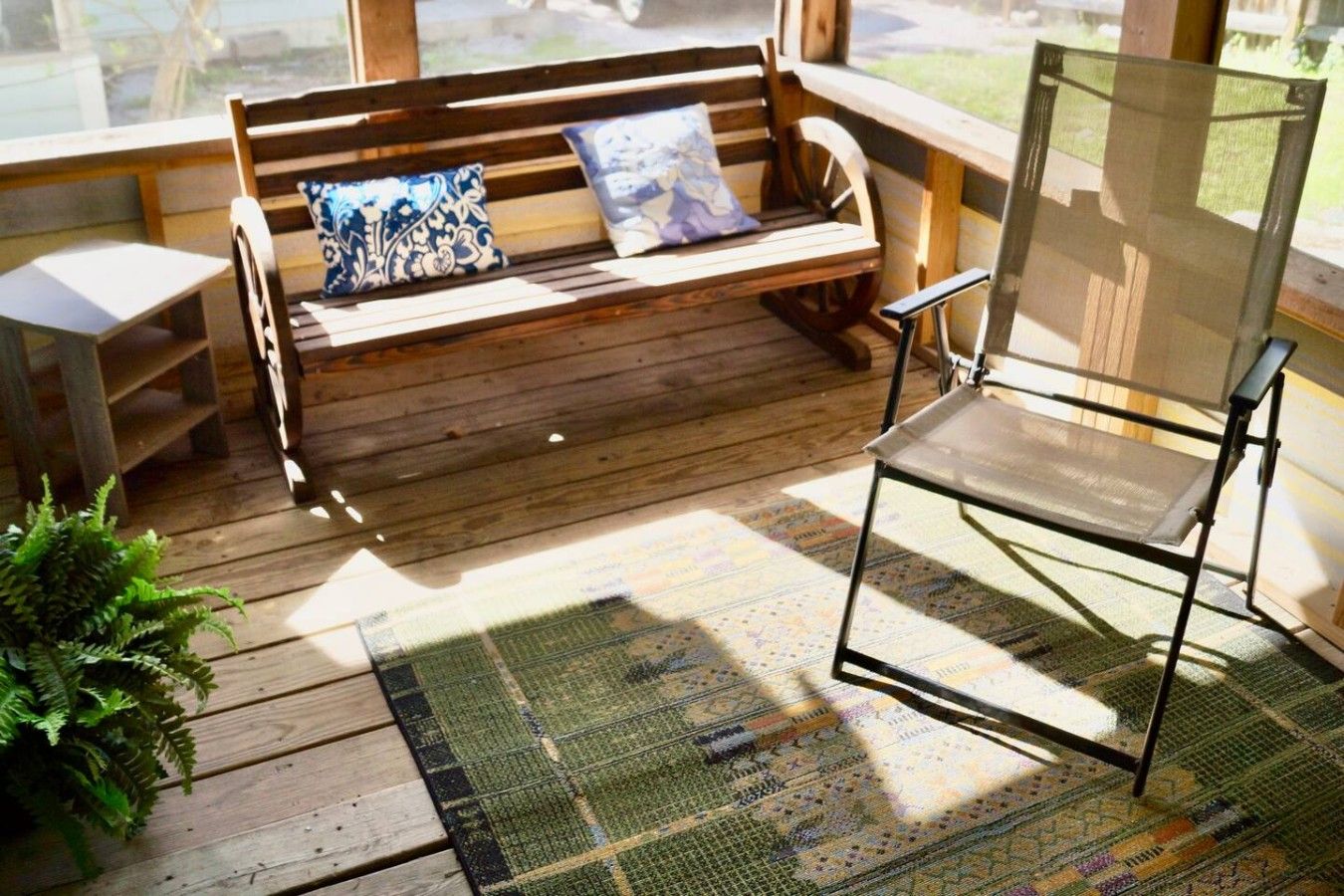 Un banco de madera con cojines, una silla de metal y una alfombra estampada sobre una terraza de madera.