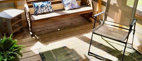 Un banco de madera con cojines, una silla de metal y una alfombra estampada sobre una terraza de madera.