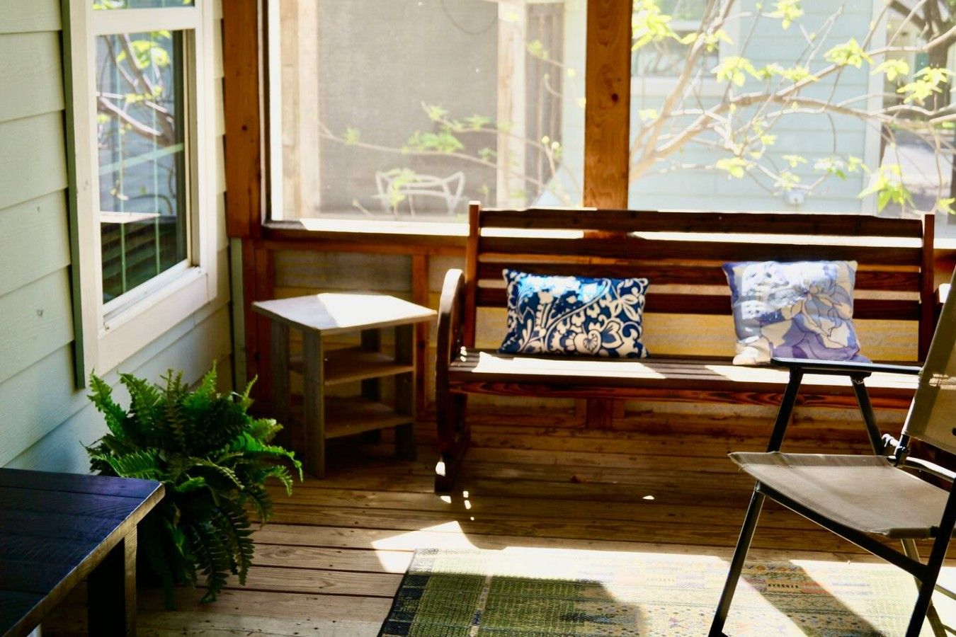 A wooden bench with a cushion, a small table, and a chair in a sunlit porch area.