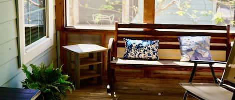 Un banc en bois avec un coussin, une petite table et une chaise, le tout installé dans un coin de véranda baigné de lumière.