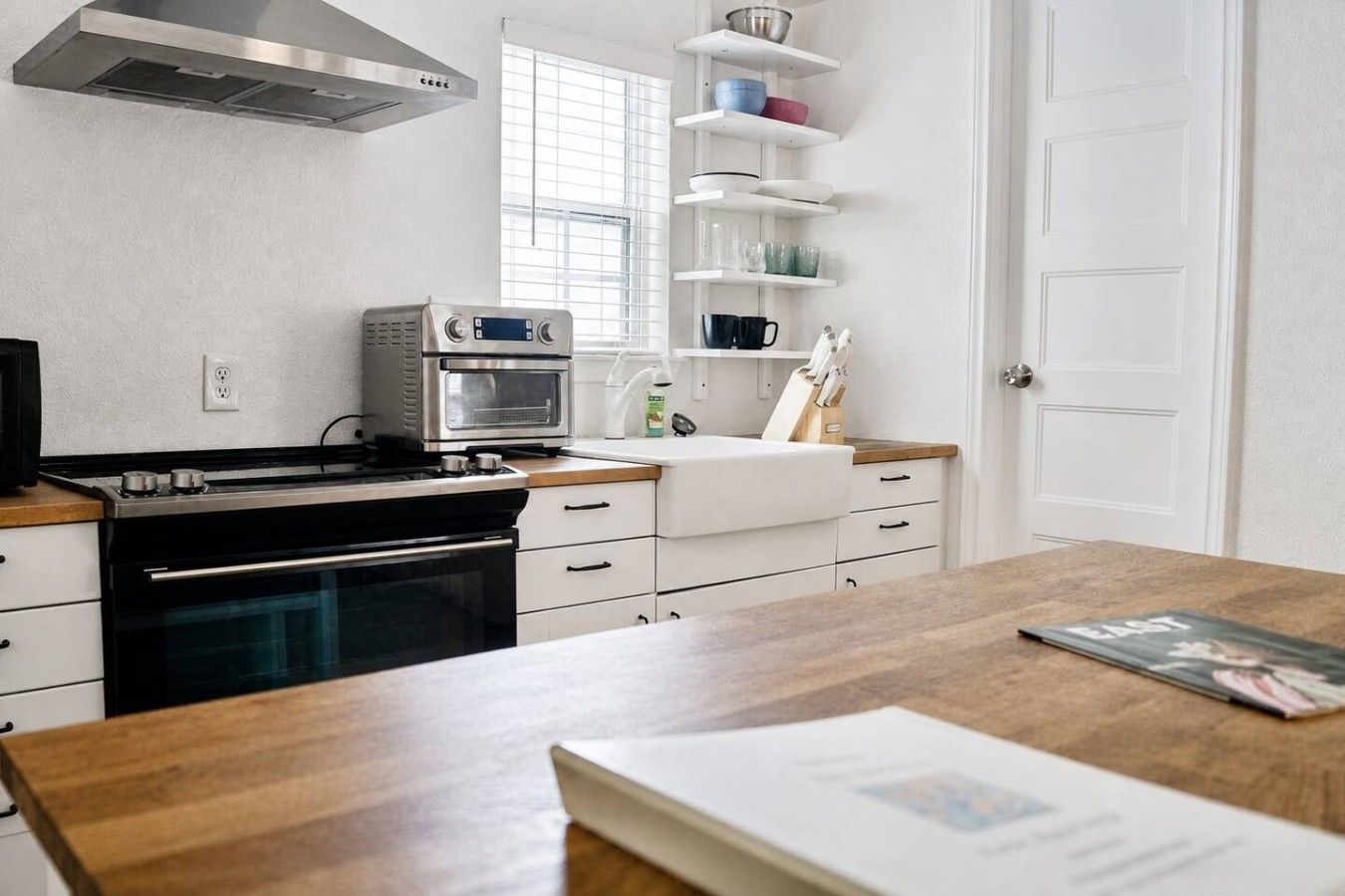 A kitchen with a stove, oven, and microwave. There is a window with blinds, a sink, and a shelf with dishes.