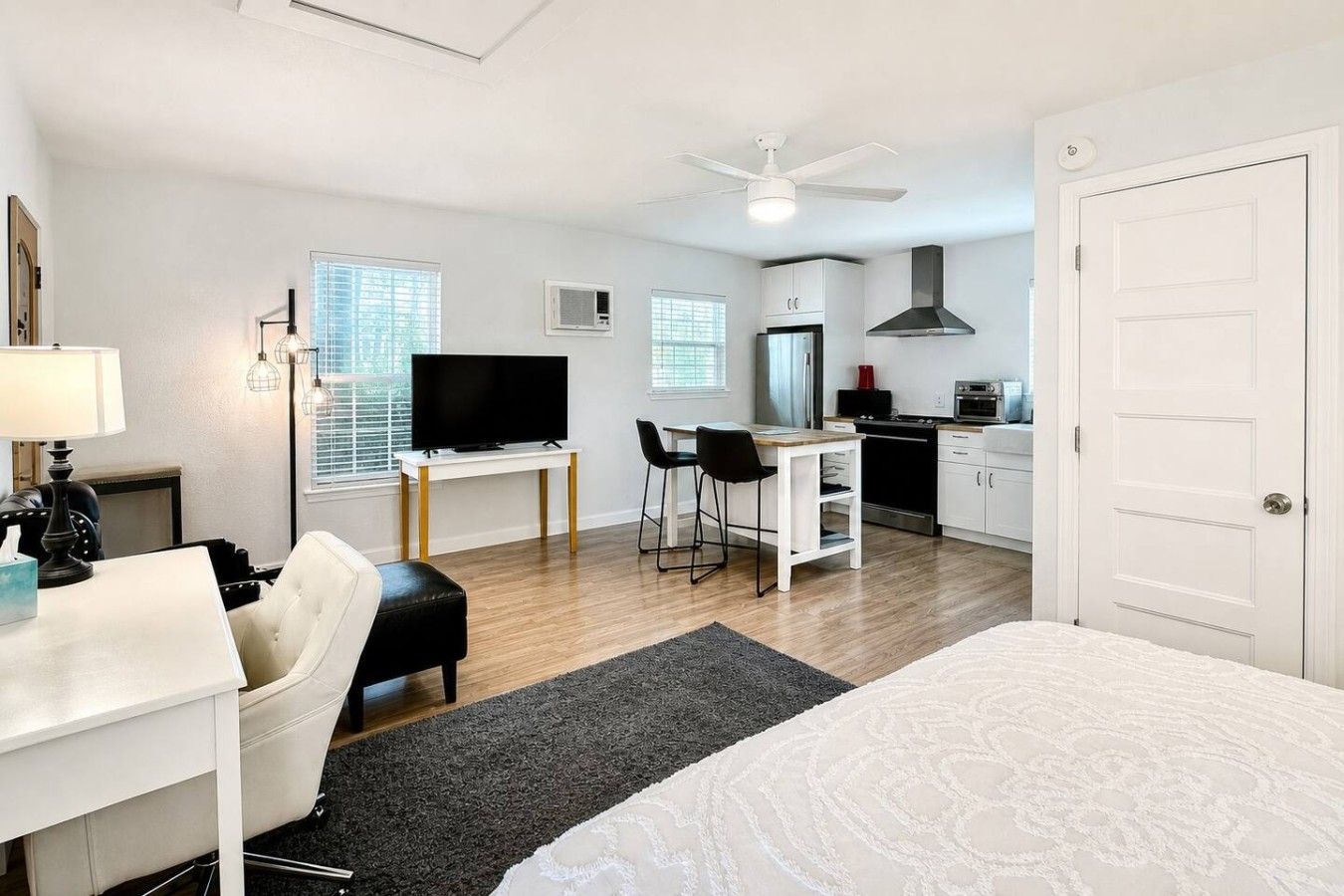 A modern hotel room with a bed, a desk, a chair, a television, a kitchenette, and a ceiling fan.