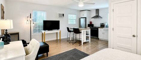 A modern hotel room with a bed, a desk, a chair, a television, a kitchenette, and a ceiling fan.