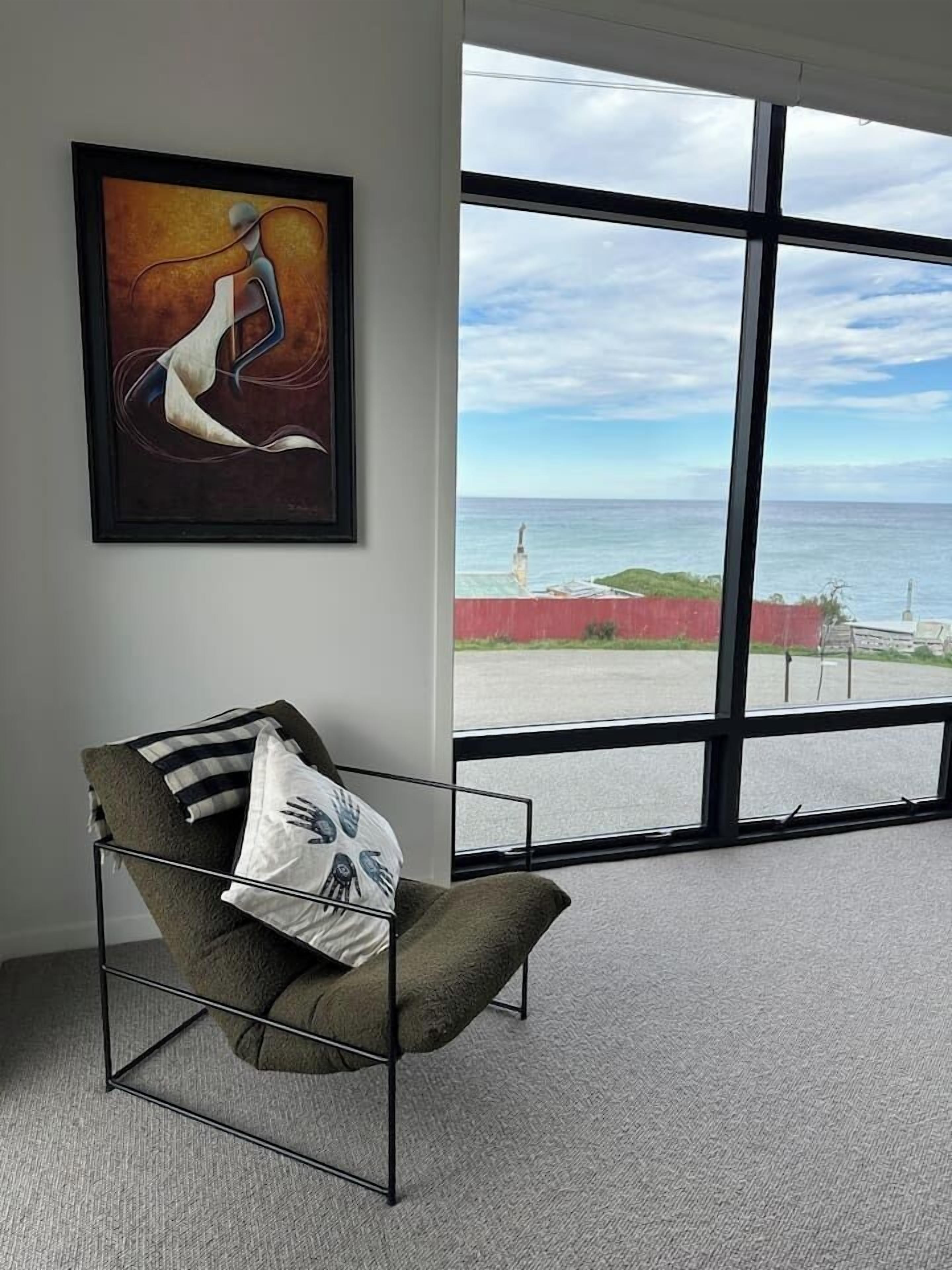 Ein Zimmer mit großem Fenster, einem gerahmten abstrakten Gemälde und einem Stuhl mit Metallgestell und Kissen.