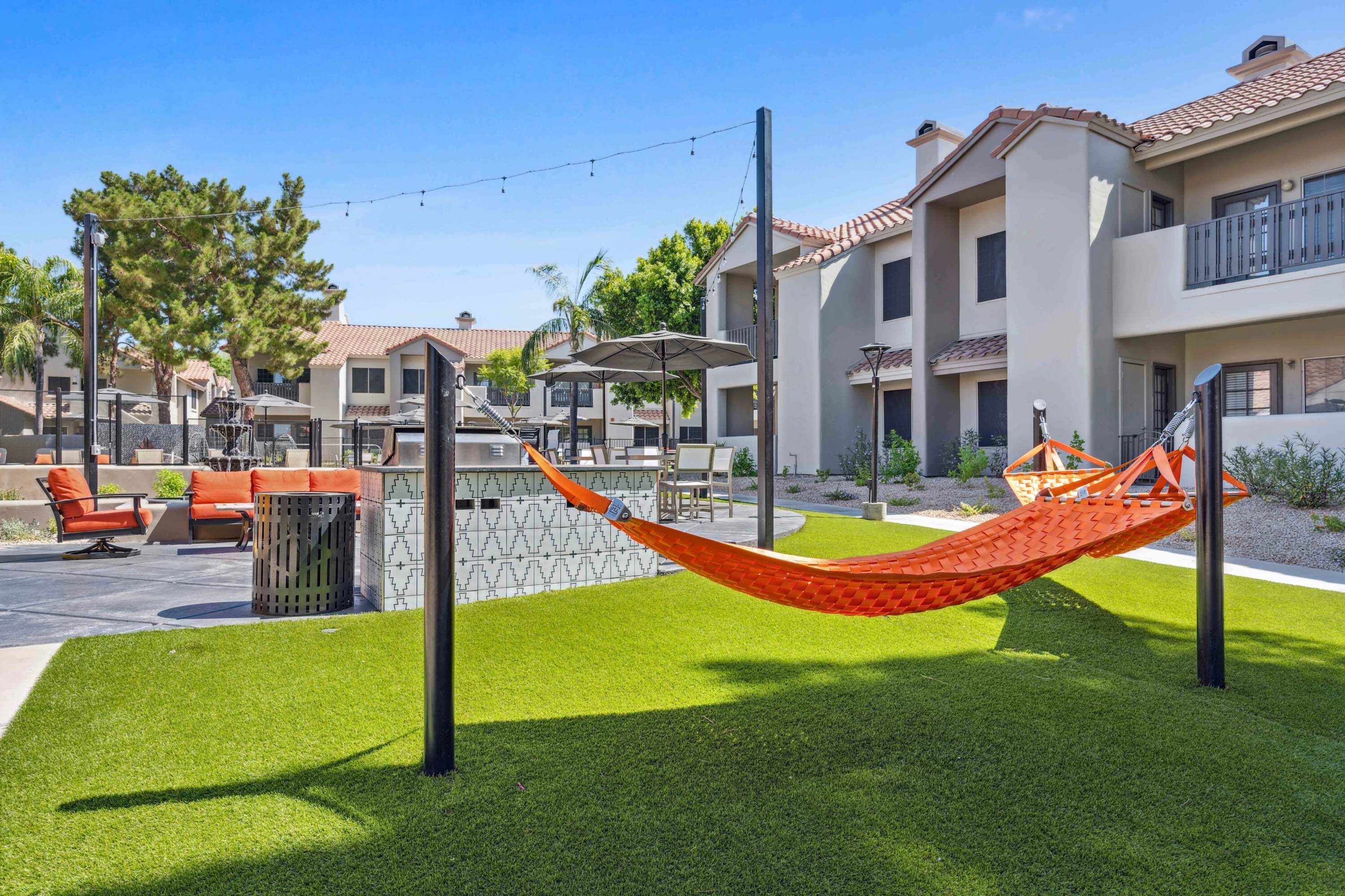 A hammock in a well-maintained grassy area with outdoor furniture and a stone wall, surrounded by modern buildings with balconies.