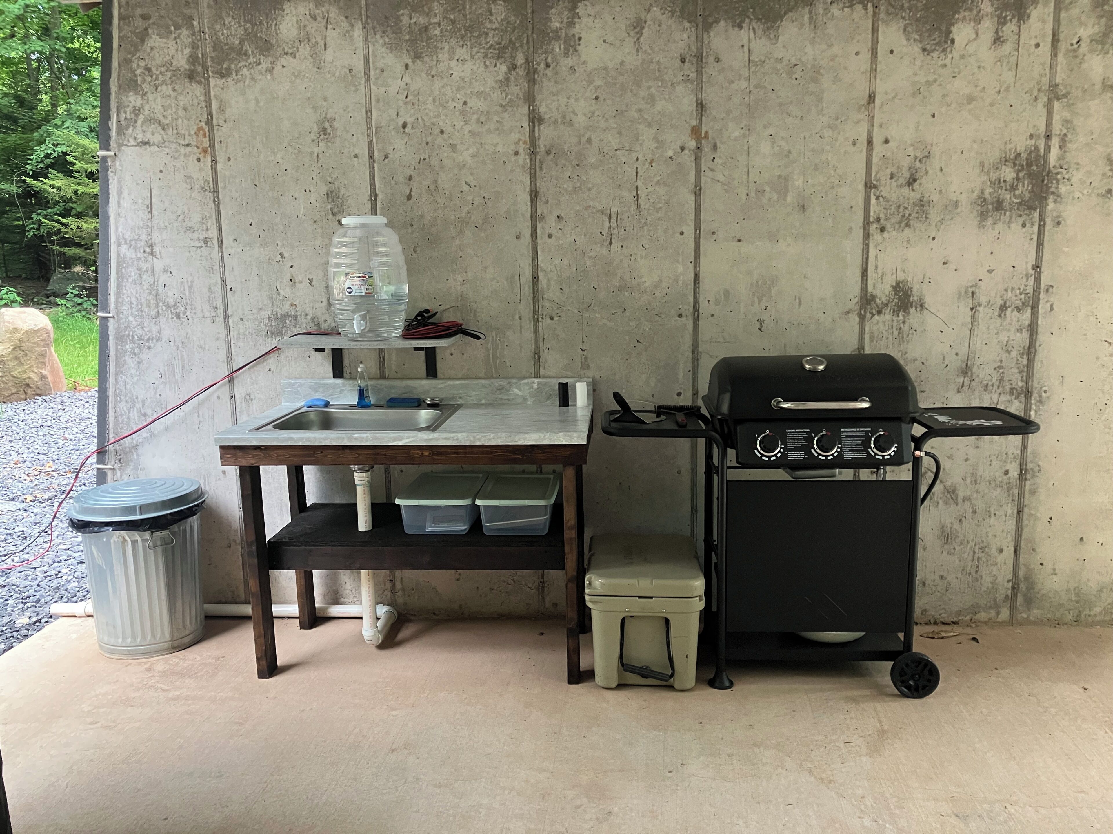 A concrete wall with a metal sink, a portable grill, and storage containers.