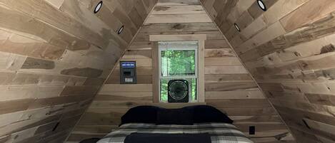 An attic bedroom with a sloped ceiling, a bed with a plaid bedspread, a window with a view of greenery, and a wall-mounted heater.