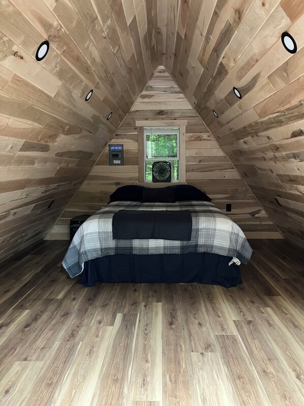 Un dormitorio en ático con techo inclinado, una cama con colcha escocesa, una ventana con vista a la vegetación y un calentador fijado a la pared.