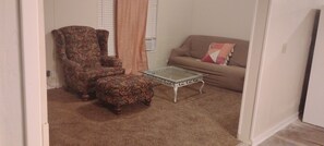 A living room with a patterned armchair, a glass-top coffee table, and a sofa with a cushion.