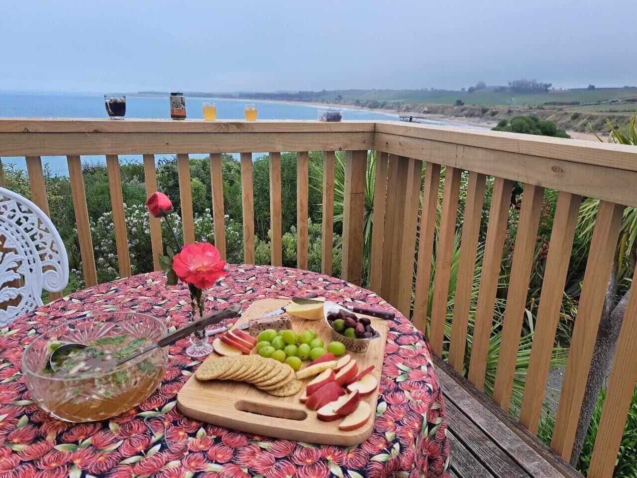 A round table with a floral tablecloth, a bowl of soup, a plate of sliced apples, grapes, cheese, and crackers, and a glass of iced tea on a balcony overlooking a beach.