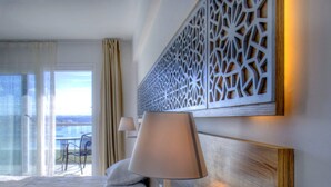Een hotelkamer met een bed, een nachtkastje met een lamp, een decoratief wandpaneel en uitzicht op zee vanaf een balkon.