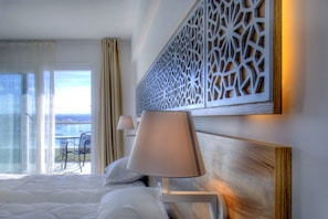 A hotel room with a bed, a nightstand with a lamp, a decorative wall panel, and a view of the sea through a balcony.