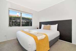 Un dormitori modern amb un llit gran, una finestra amb vista a vegetació i un edifici, i un capçal de llit fosc.
