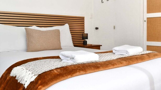 Uma cama bem arrumada com um cobertor marrom e duas toalhas brancas dobradas, uma cabeceira de madeira, um abajur e uma porta com puxador.