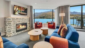A modern living room with a stone fireplace, comfortable seating, and a large window offering a scenic view. - Postcard Views & Pure Comfort: Your Lakeside Haven (Queenstown)