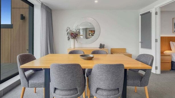 A modern dining area with a wooden table and grey upholstered chairs, a round mirror on the wall, and a view of a balcony. - Postcard Views & Pure Comfort: Your Lakeside Haven (Queenstown)