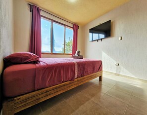 A single bed with a wooden frame and a red bedspread in a room with a large window, a flat-screen TV mounted on the wall, and a tiled floor.