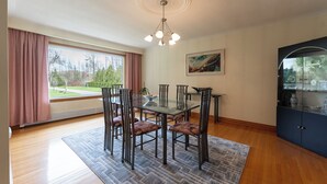 Spiseområde med et glasbord, metalstole, et mønstret tæppe, en loftlampe og et vindue med gardiner.