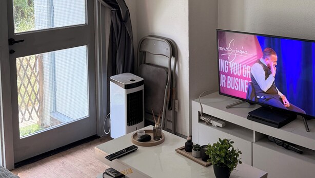 Une pièce avec une grande fenêtre, un téléviseur, un bureau avec un clavier, une chaise et une petite plante.