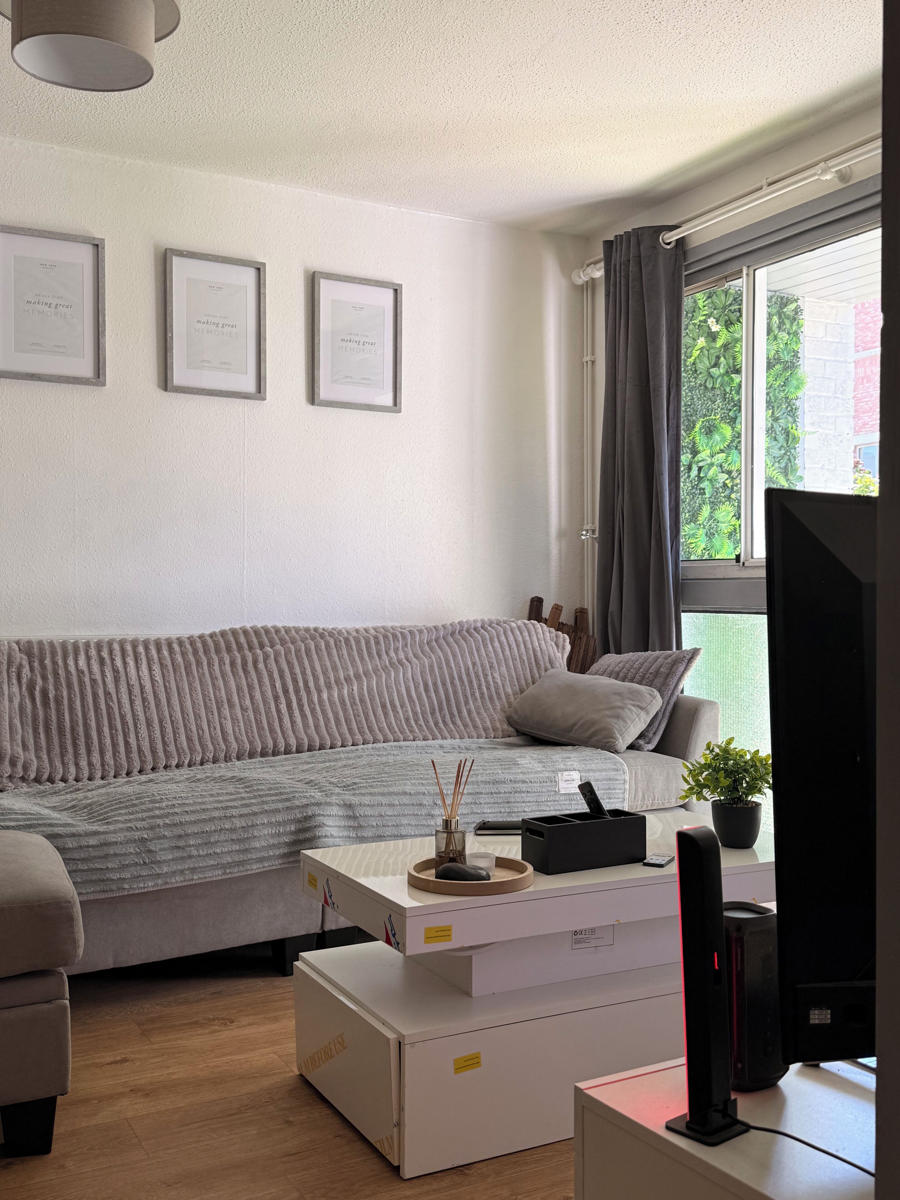 A modern living room with a grey sofa, a white coffee table, and a television. There are framed certificates on the wall and a window with curtains.